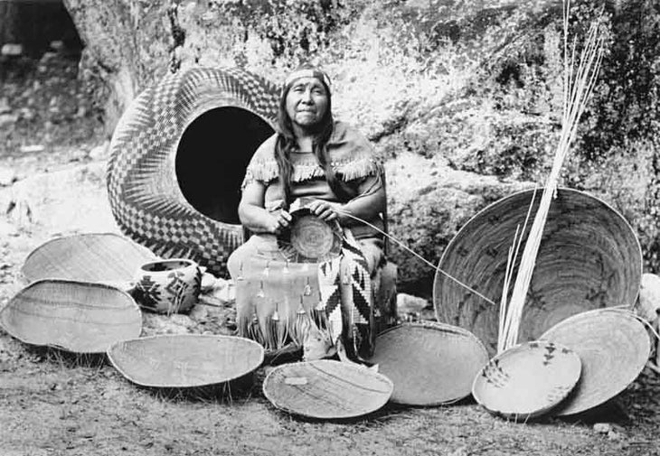 Lucy Telles, also known as Pa-ma-has (1885-1955) working on a basket, seated amidst ten other baskets, possibly all made by her.